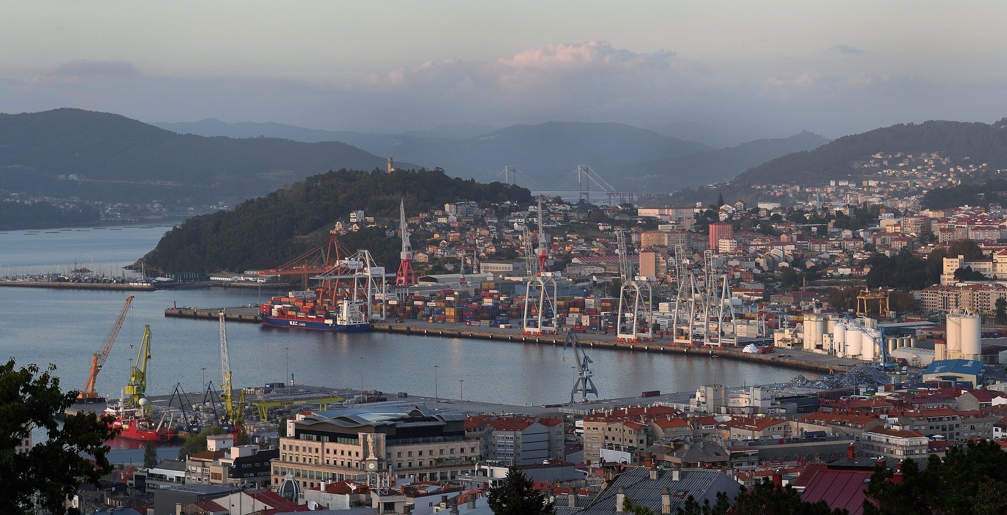 Vigo at dusk on a late-September evening, seen from high above the city centre: the long quay of the Guixar container terminal curves along the inner Ría de Vigo, lined with white gantry cranes and a container ship, yellow and orange harbour cranes rise in front of the rooftops of the centre, the wooded hill of A Guía crowned by a small tower stands on the left above a marina, and far beyond, between layers of blue-grey mountains, the cable-stayed Rande bridge spans the ría under a soft pink-tinged sky