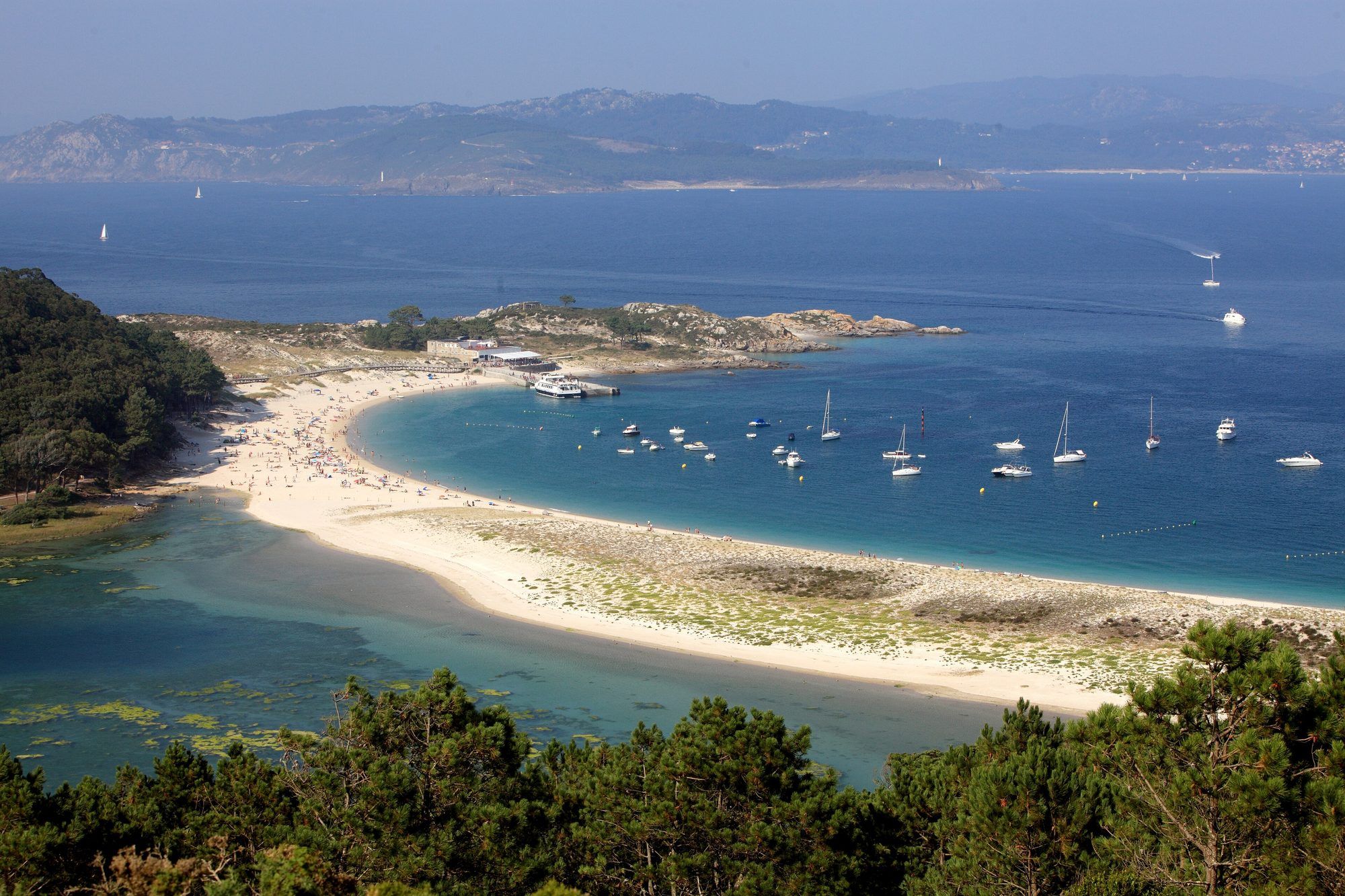 Praia de Rodas on the Cíes Islands seen from the hillside above on a hazy summer day: a long arc of white sand joins two of the islands, with clear turquoise water dotted with anchored sailing and motor boats on the right and the shallow green lagoon on the left, a ferry is tied up at the landing pier at the far end of the beach below a rocky point, pine trees fill the foreground, and across the deep-blue mouth of the Ría de Vigo rise the hills of the Morrazo peninsula